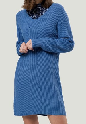 Femme portant une robe pull en maille côtelée bleue à col en V sur un haut à col montant à motif, bras croisés à la taille, sur un fond uni.