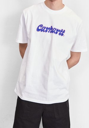 Osoba ubrana w biały T-shirt z krótkim rękawem z niebieskim napisem "Carhartt" i czarne spodnie, stoi z rękami za plecami na tle jednolitej ściany.