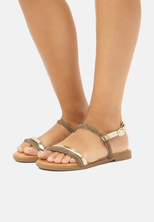 Sandalias planas doradas y marrones con tiras, dos bandas anchas y cierre de hebilla; suela plana con un diseño texturizado en las tiras.