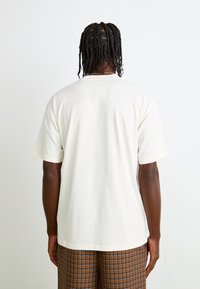 Light cream cotton t-shirt with a relaxed fit, short sleeves, and a round neckline, paired with plaid brown and blue pants.