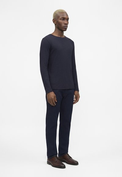 Άνδρας με κοντά ξανθά μαλλιά στέκεται, φορώντας μπλε σκούρο πουκάμισο μακρυμάνικο, μπλε σκούρο παντελόνι και καφέ παπούτσια επίσημα, μπροστά σε απλό φωτεινό φόντο.