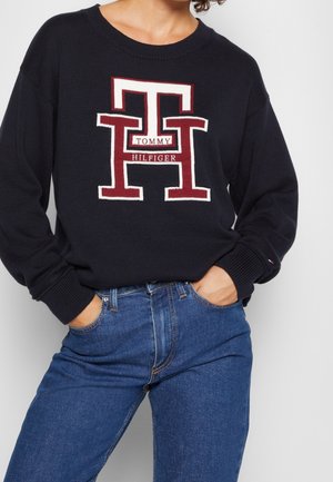 Osoba ubrana w czarny sweter z dużym czerwono-białym logo "TH" i napisem "Tommy Hilfiger", w połączeniu z niebieskimi dżinsami, z rękami w kieszeniach.