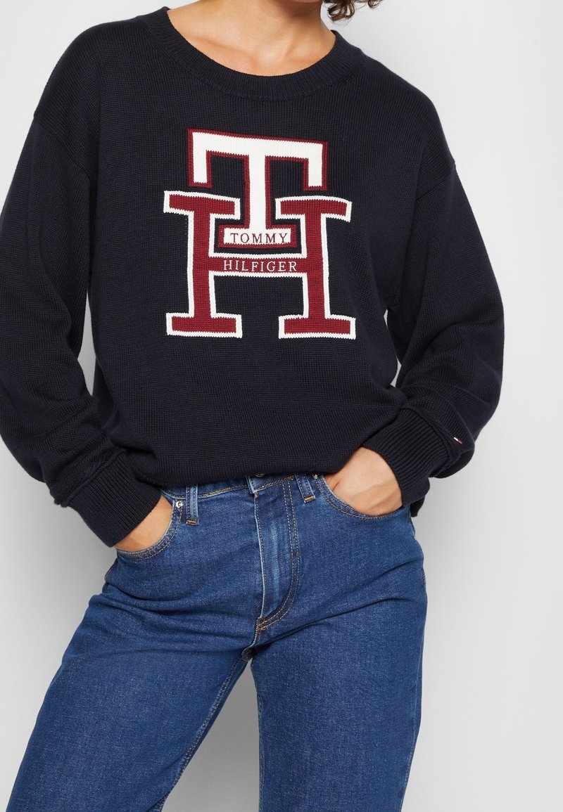 Personne portant un pull noir avec un grand logo "TH" rouge et blanc ainsi que le texte "Tommy Hilfiger", associé à un jean en denim bleu, mains dans les poches.