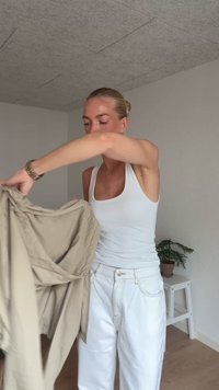 Beige vnější oděv držený osobou, s volným, splývavým designem. Pod ním bílé tílko a bílé kalhoty s vysokým pasem.