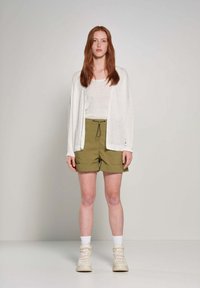 Cardigan leggero di crema indossato sopra una maglietta bianca aderente e pantaloni corti verde oliva. L'outfit è abbinato a sneaker beige alte.