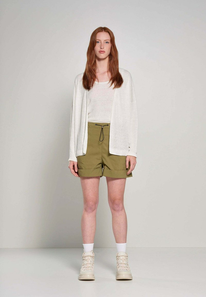Cardigan leggero di crema indossato sopra una maglietta bianca aderente e pantaloni corti verde oliva. L'outfit è abbinato a sneaker beige alte.