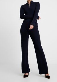 Marine ribbel jumpsuit met een ritssluiting bij de hals, lange mouwen en uitlopende benen, gecombineerd met donkere, gestructureerde hakken. Zacht, aansluitend materiaal.
