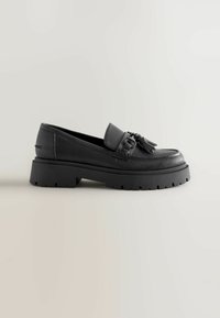 Zapato negro de cuero tipo loafer con acabado texturizado, detalle de borlas ajustable y suela de goma gruesa con un patrón de surcos.