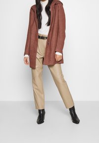 Abrigo con capucha color óxido, tejido texturizado, pantalones rectos beige, botines negros, complementados con un cinturón negro con acentos metálicos.