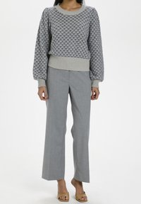 Heller grauer Strickpullover mit strukturiertem Muster; kombiniert mit leichten, gerade geschnittenen grauen Hosen. Braune Sandalen mit offenen Zehen runden den Look ab.