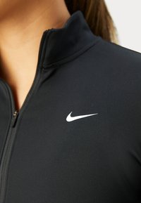 Musta urheilu-zip-up-takki, jossa on korkea kaulus ja valkoinen Nike-logo vasemmalla ylhäällä. Kangas näyttää sileältä ja istuvalta.