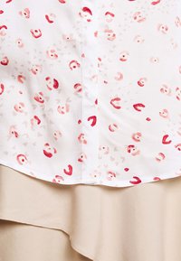 Blouse blanche avec des taches et des tourbillons irréguliers rouges et rose clair, dotée d'une fermeture à boutons sur le devant et d'une couche inférieure beige à volants.