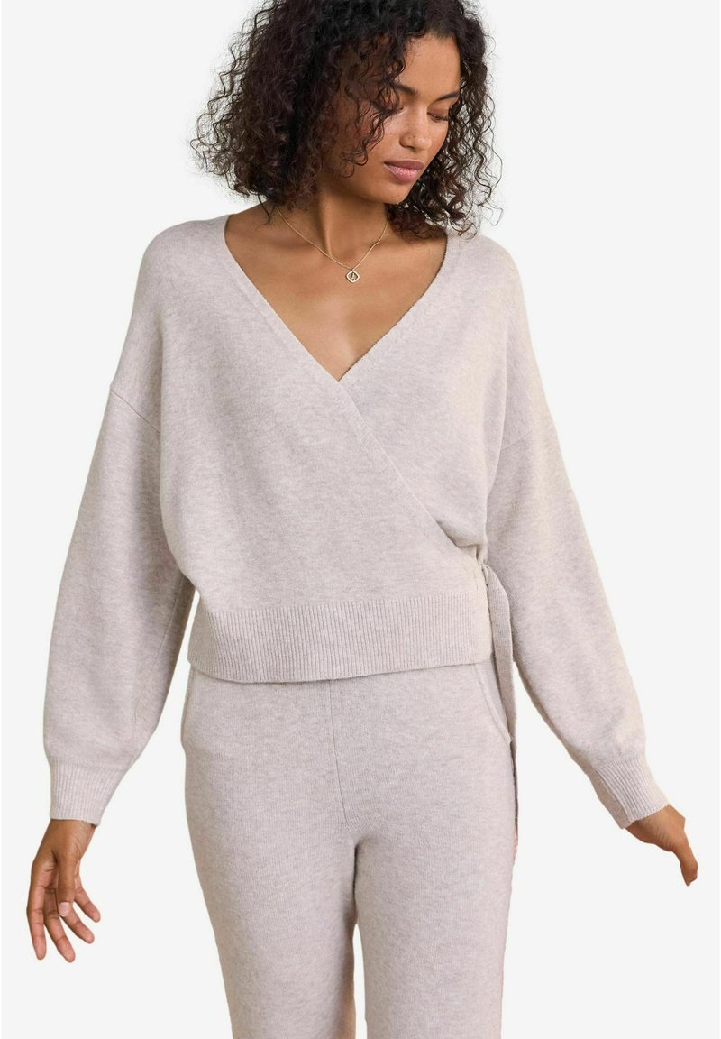 Svetra v svetlo sivi barvi, zavita, iz mehkega pletenega materiala, z V-izrezom, oversized rokavi in ribastimi detajli na robu. Ustvarjeno v kombinaciji z usklajenimi hlačami.