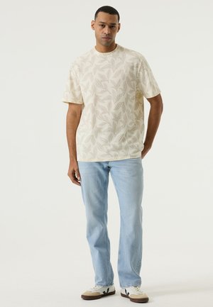 Mann steht und trägt ein cremefarbenes kurzärmeliges Hemd mit Blattmuster, hellblaue Jeans und weiße Sneakers mit schwarzen Details.