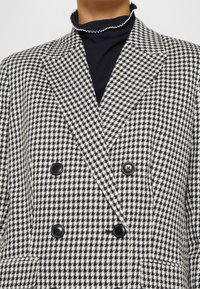 Zwart-witte houndstooth dubbelzijdige blazer met zwarte knopen, gedragen over een zwart top met een ruche kraag.