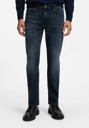 Mann trägt dunkelblaue Slim-Fit-Jeans, schwarze Lederschuhe und einen marineblauen Pullover und steht vor einem einfarbigen Hintergrund.