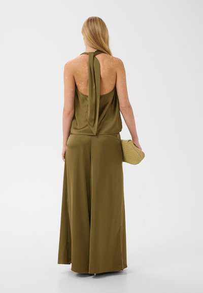Femme aux longs cheveux blonds portant un haut dos-nu vert olive et un pantalon à jambes larges, tenant une pochette beige tressée, tournée dos à la caméra sur fond blanc.