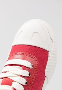 Baskets en toile rouge avec un capuchon en caoutchouc blanc, des lacets contrastés et un tissu texturé. Présente un détail en cuir lisse le long des bords.