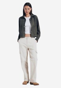 Krótka kurtka w oliwkowo-zielonym kolorze z dwoma przednimi kieszeniami, zestawiona z beżowymi spodniami cargo z bocznymi kieszeniami. Całość dopełniają brązowe buty na obcasie.