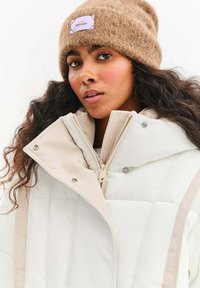 Kurtka puchowa w kolorze ecru z prążkowanym kołnierzem, zamkiem błyskawicznym i zatrzaskami. Brązowa dziana czapka z detalem w postaci metki.