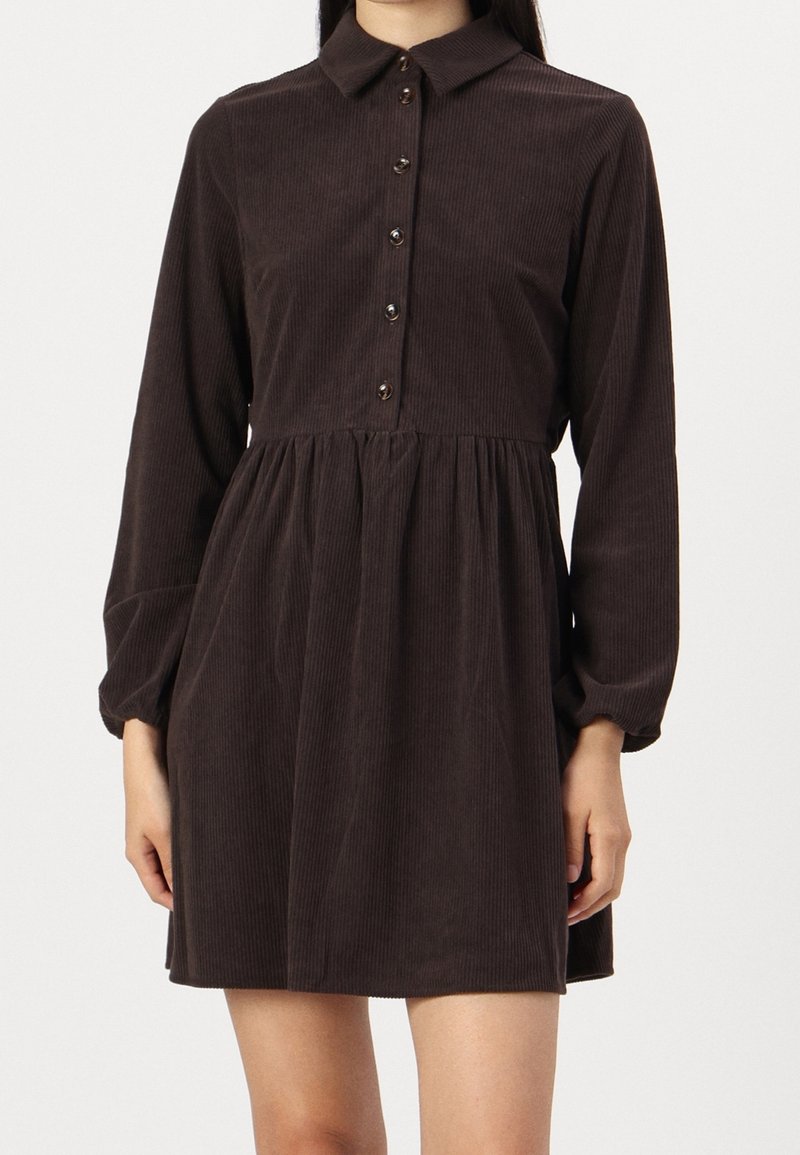 Femme portant une robe en velours côtelé marron foncé avec des manches longues, un col et cinq boutons sur le devant.