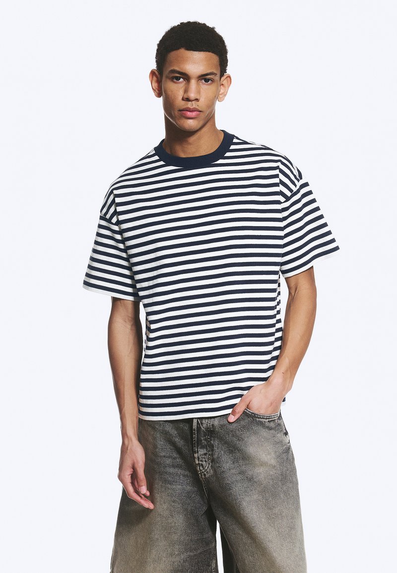 Jeune homme portant un t-shirt à manches courtes rayé noir et blanc et un jean noir délavé et ample, debout avec une main dans la poche.
