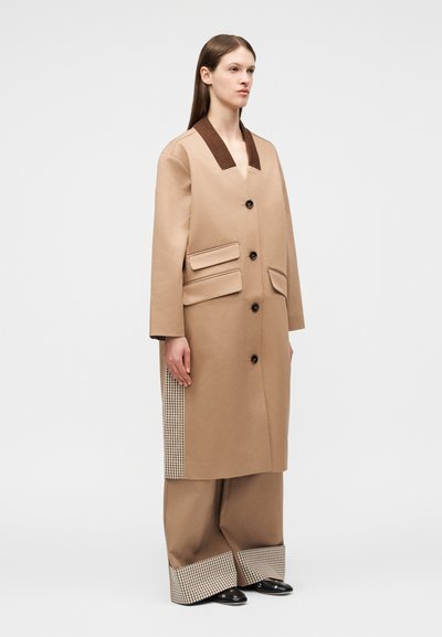 Γυναίκα που φοράει μια μπεζ μακριά καμπαρντίνα με καφέ γιακά και καρό πλαϊνές πινελιές, ταιριαστά φαρδιά παντελόνια και μαύρα παπούτσια, όρθια πλαγίως.