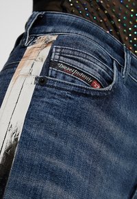 Gros plan sur un jean en denim bleu avec une bande métallique argentée et une étiquette de marque Diesel Industry sur la petite poche avant.