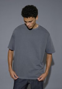 T-shirt oversize in cotone grigio con colletto a girocollo, maniche corte e vestibilità rilassata, abbinato a jeans scuri. Design semplice e monocolore.