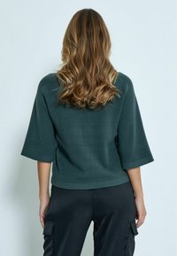 Maglione verde scuro, con texture, a maniche ¾ e vestibilità rilassata, abbinato a pantaloni neri, lucidi e larghi. Nessun motivo particolare o dettagli in metallo.