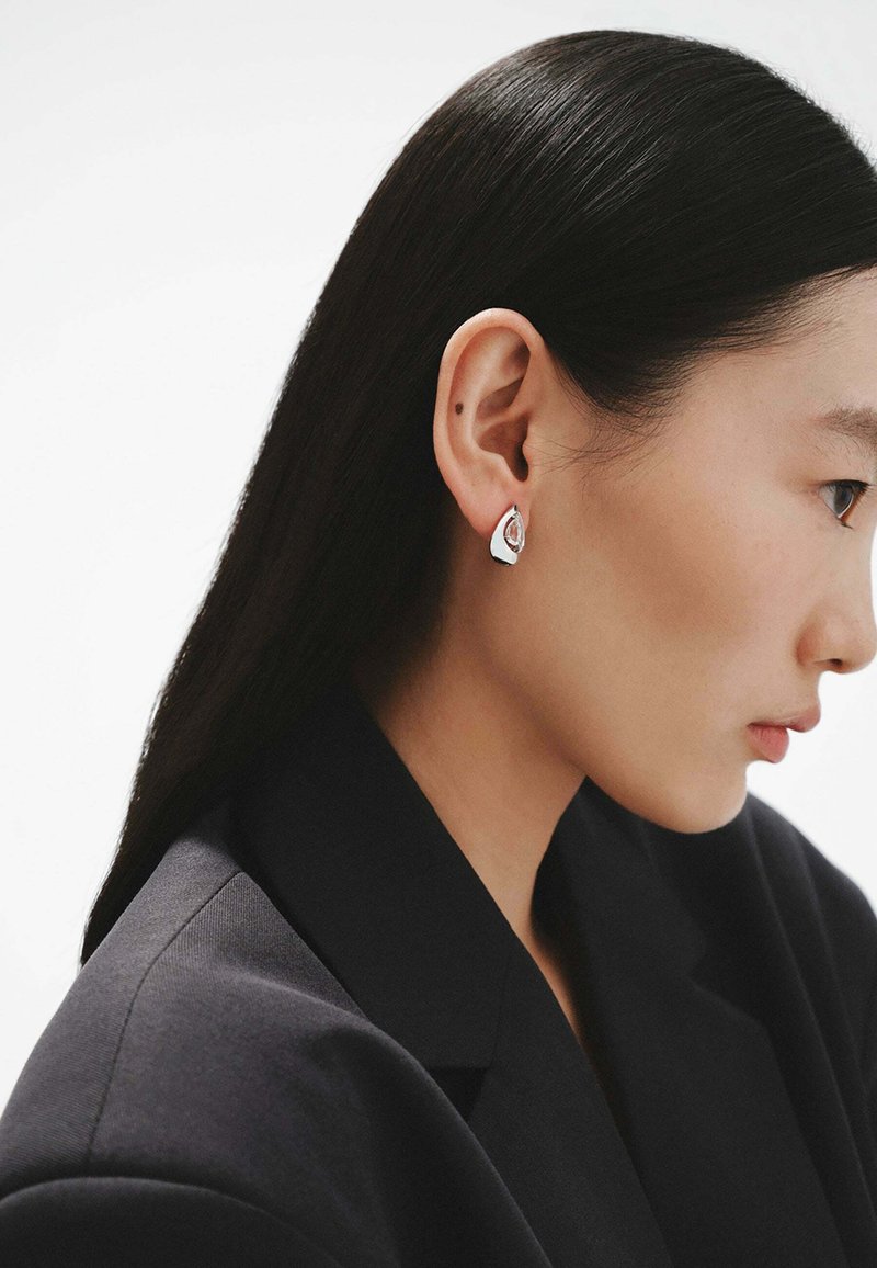 Pendientes en forma de lágrima de plata con un diseño en capas, acabado brillante y un corte central, llevados por una modelo con el cabello lacio y oscuro.