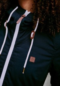 Czarna kurtka z kapturem z białymi sznurkami, z brązowymi skórzanymi akcentami i metką marki. Zapięcie na suwak z przodu. Gładka faktura.