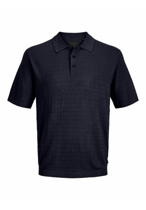 Nastolatek uśmiecha się, ubrany w czarną koszulkę polo z krótkim rękawem, podwinięte jeansowe szorty, białe skarpetki i czarno-białe trampki, stoi na jednolitym tle.