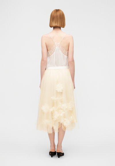Ένα αέρινο φόρεμα σε κρεμ απόχρωση με εφαρμοστό τοπ, λεπτές τιράντες και μια ροή, στρωματοποιημένη φούστα από τούλι διακοσμημένη με εφαρμογές λουλουδιών.