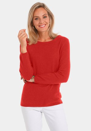 Pull rouge à manches longues et décolleté large, fabriqué en matière tricot douce. Porté avec un pantalon blanc. Design simple, sans motifs ni accents.