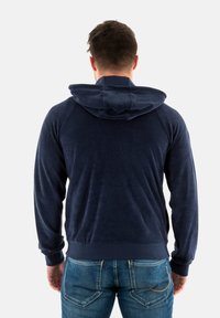 Navyblauwe hoodie van zacht, gestructureerd materiaal; beschikt over een kangoeroezak en geribde manchetten. Draagt in combinatie met denim jeans.