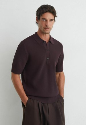 Homme aux cheveux courts et foncés porte un polo tricoté violet foncé à manches courtes et un pantalon foncé, debout devant un mur clair uni.
