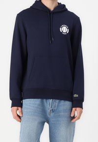 Námořnická modrá mikina s kapucí vyrobená z bavlněné směsi. Má přední kapsu, logo Lacoste na hrudi a žebrované manžety a lem.