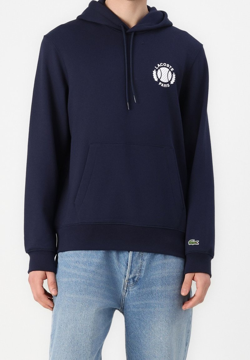 Námořnická modrá mikina s kapucí vyrobená z bavlněné směsi. Má přední kapsu, logo Lacoste na hrudi a žebrované manžety a lem.