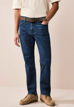 Donkerblauwe denim jeans met een rechte snit, voorzien van subtiele vervaagde accenten, gecombineerd met een bruine leren riem en beige suède schoenen.
