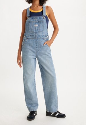 Salopette en denim bleu clair avec bretelles ajustables, poche avant et boutons sur les côtés, assortie à un débardeur bleu marine et des baskets noires.