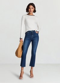 Long-sleeve white top with navy neckline, paired with high-waisted dark blue jeans and beige bag. Model wears light-colored sandals.