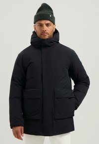 Černá vatovaná bunda s vysokým límcem, velkými předními kapsami a zipovým uzavřením. Nosená s tmavě zelenou pletenou čepicí.