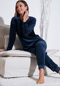Navy sininen samettipyjama-asuste, jossa on pitkät hihat ja kapeat housut, pehmeällä tekstuurilla ja ribatuilla hihansuilla, tyylitelty vaalealla sohvall