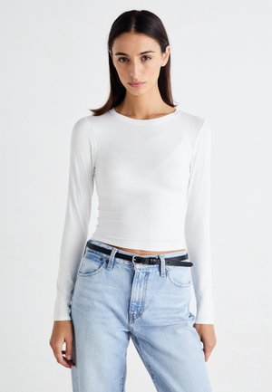 Femme portant un haut blanc ajusté à manches longues, rentré dans un jean taille haute bleu clair avec une ceinture noire, debout devant un fond uni.
