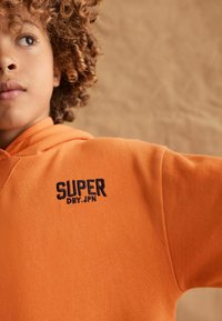 Mlada osoba s kovrčavom kosom koja nosi narančastu hoodicu s izvezenim natpisom "SUPER DRY.JPN" na prsima, gleda u stranu.