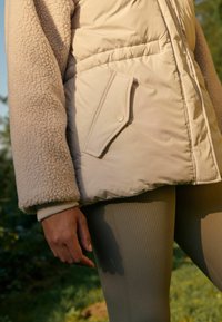 Doudoune beige avec des manches en polaire et une poche à rabat boutonnée, accompagnée de leggings olive côtelés. Texture douce et palette de couleurs chaleureuse.