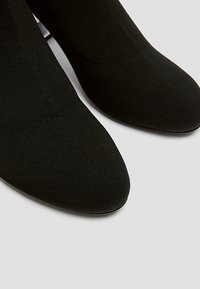 Bottes chevilles en tricot noir avec un design à bout pointu, présentant une texture lisse et une silhouette ajustée, sur un fond neutre.