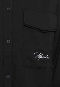 Black button-up shirt made of woven fabric, featuring a left chest pocket and embroidered "Pegador" logo in white.