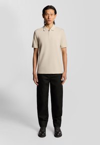 Beige zip-up polo skjorta med korta ärmar, i kombination med svarta löst sittande byxor och svarta mönstrade skor. Slät tygtextur, minimalistisk design.